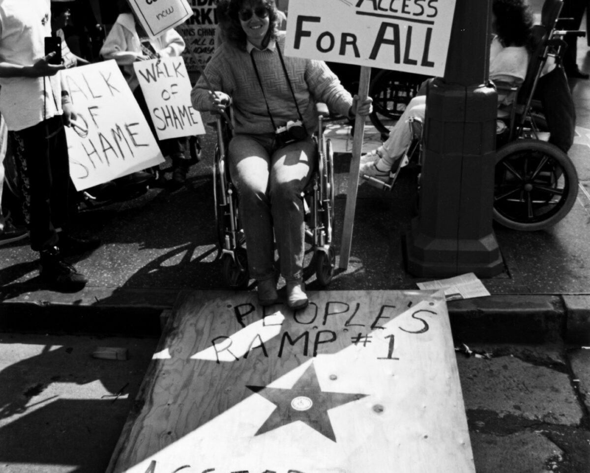 A black-and-white photo of people protesting. Some are in wheelchairs and some are standing. One person is in a wheelchair and holding a sign that says, “Build = Access For All,” and another sign that says, “Curb Cuts Now.” They are using a ramp made out of a piece of wood, that reads, “People’s Ramp #1. Access Now.” The ramp has a star with the international symbol of access in the middle of it.