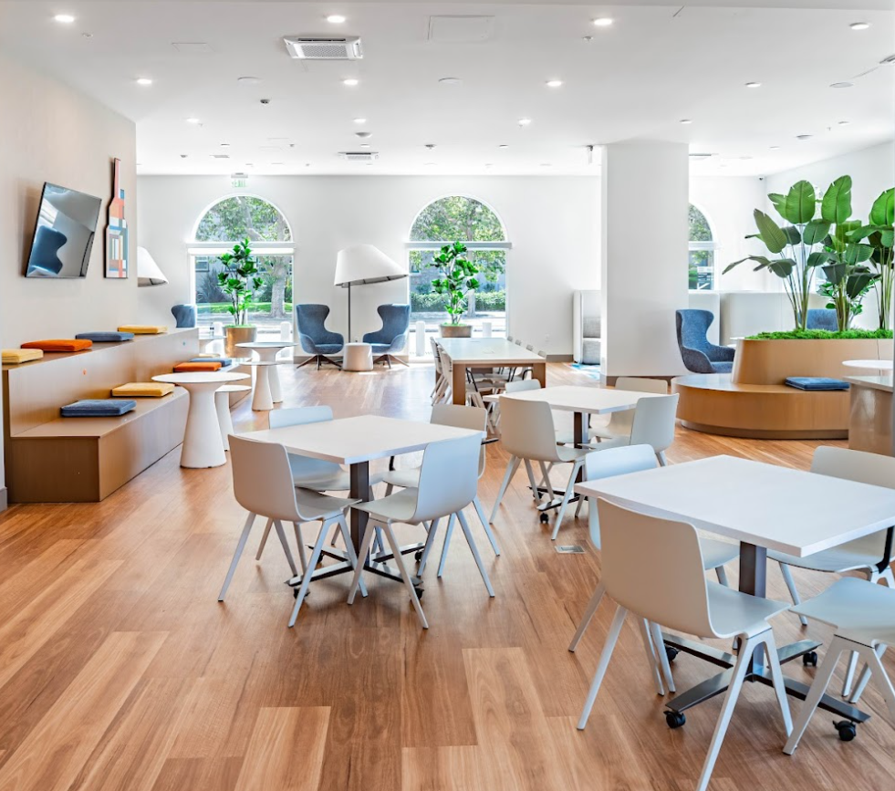 The Kelsey Ayer Station lobby. Small tables with chairs, a banquet table, colorful pillows on bench seating, a TV, and large plants are seen around the room. Large windows let in ample sunlight. 