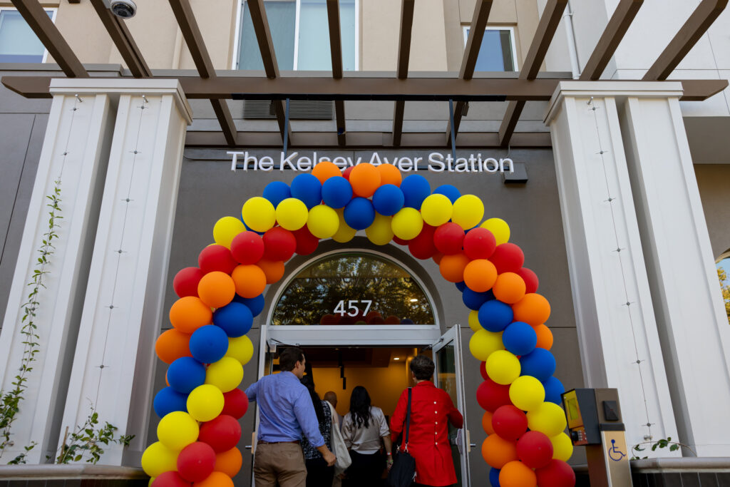 People walk in the front entrance of The Kelsey Ayer Station. A yellow, orange, red, and blue balloon arch frames the doorway and a sign with the building’s name hangs above it. 