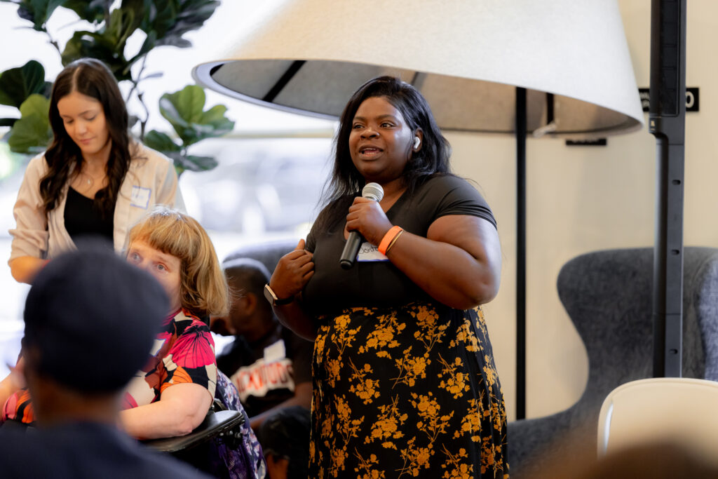 A group of residents with and without disabilities sit on a panel. A Black woman with long, straight hair is speaking into a microphone. She is wearing a black T-shirt and a black flower-patterned skirt. 