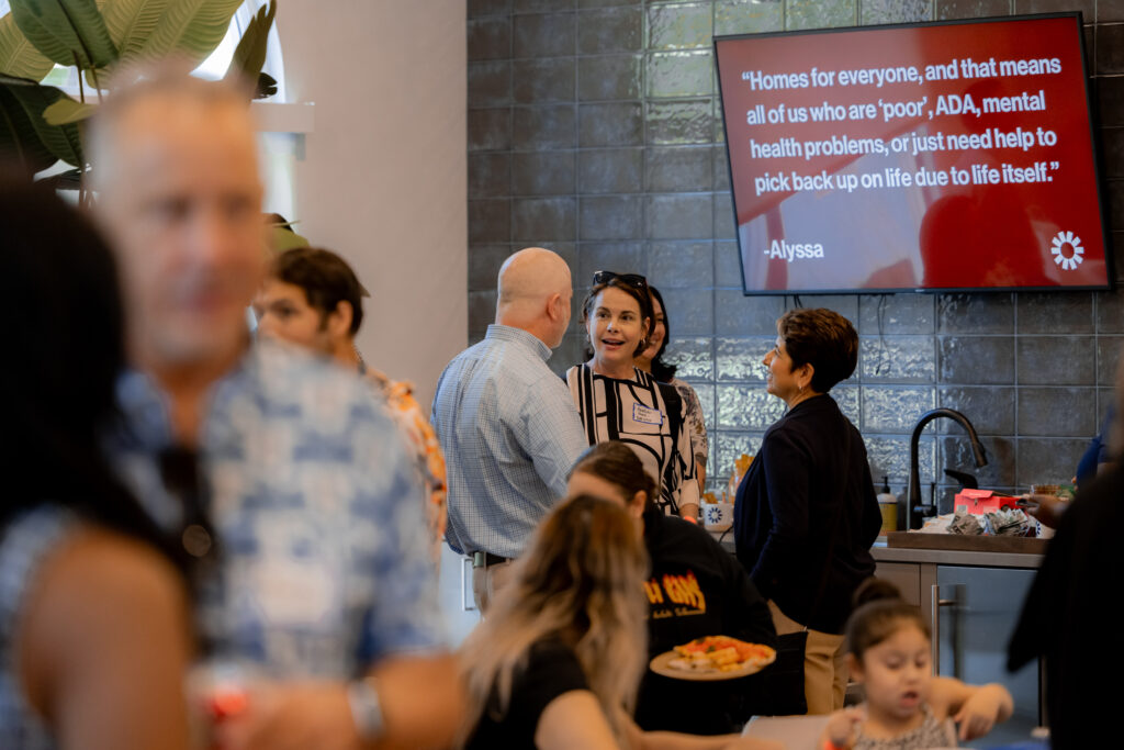 People talk in front of a TV screen displaying a quote about what inclusive homes mean. Other people socialize around the group but the camera stylistically has them out of focus. 