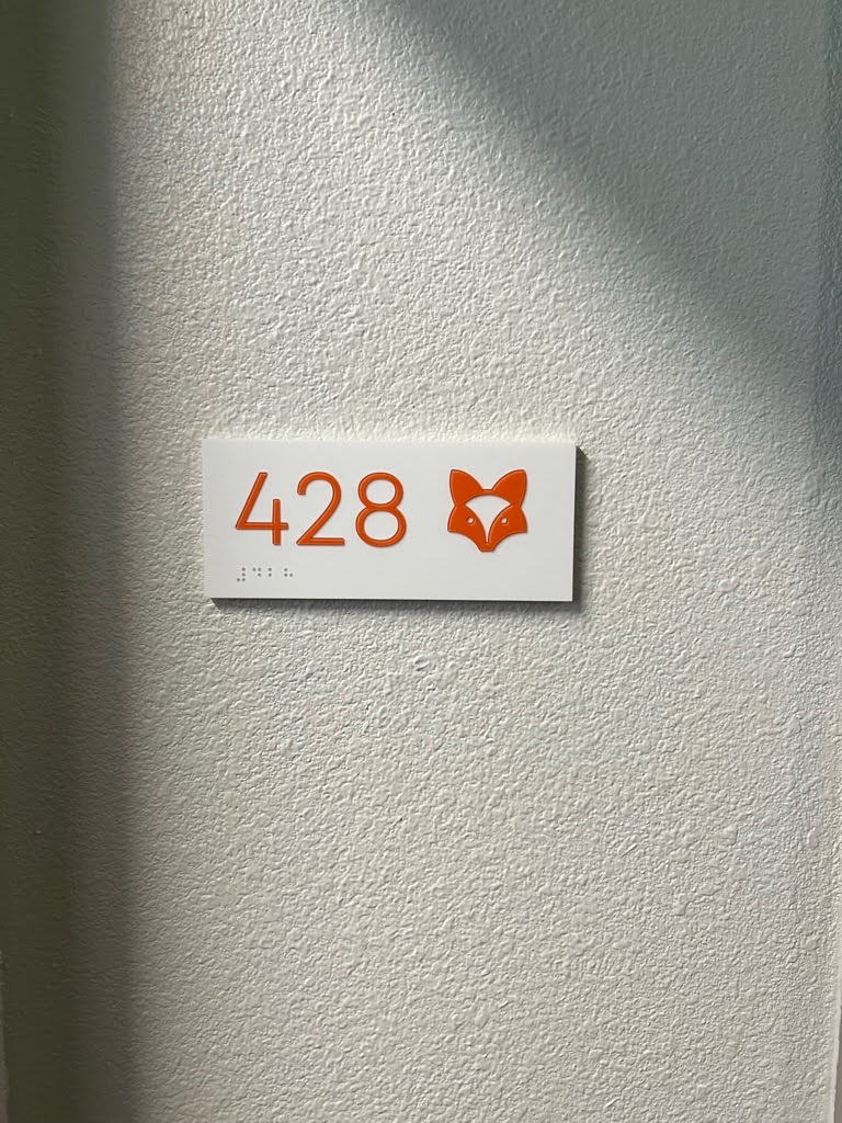 An apartment unit sign for apartment 428, written in orange text with a fox illustration next to it. The unit number is also listed in Braille below. 