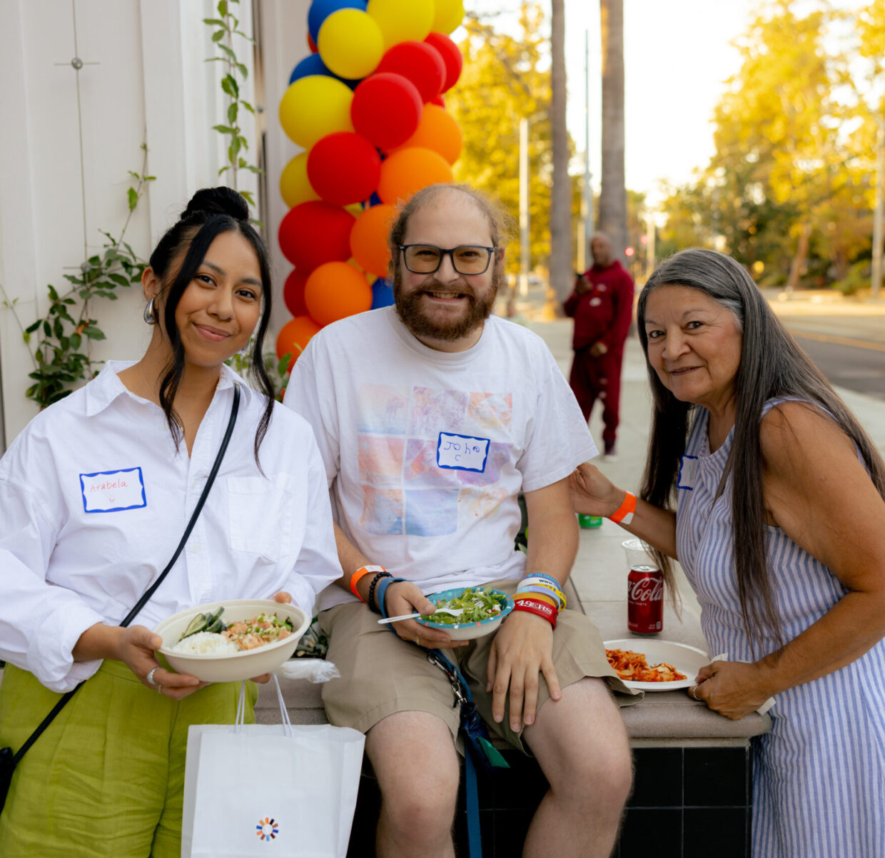 Three people pose and smile in front of The Kelsey Ayer Station during the community’s grand opening celebration. They all are holding plates of food and a colorful balloon arch is visible behind them.