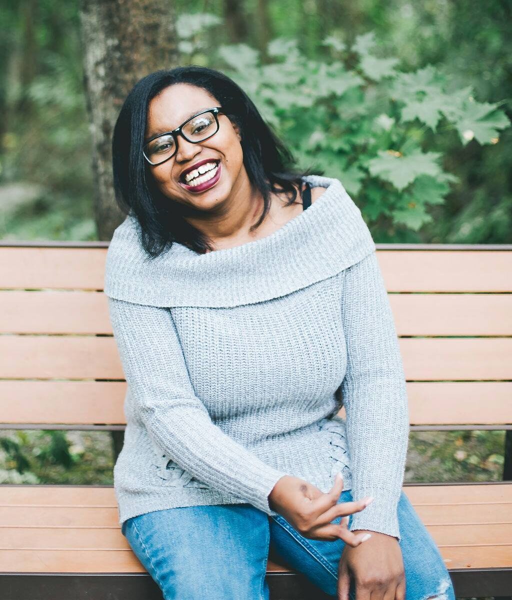 A photo of Keah Brown. A black woman with shoulder length hair wearing glasses and blue sweater sitting on a bench.