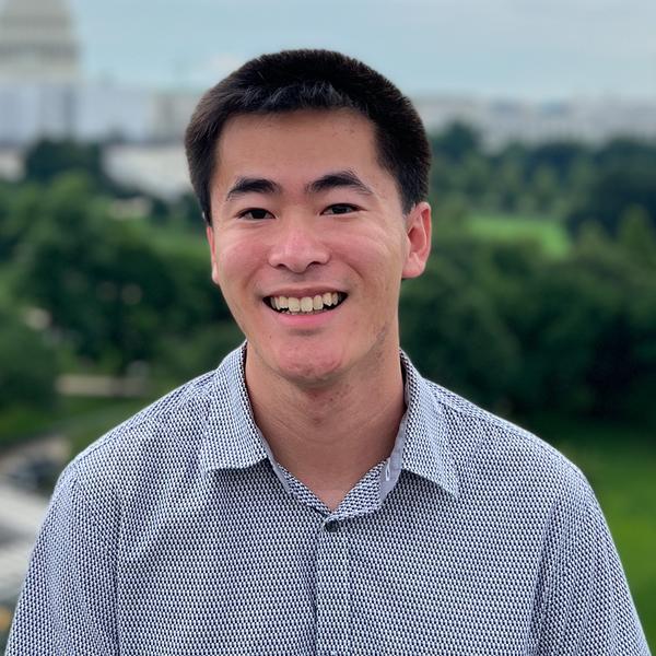 A photo of Justin Tsang and asian person with short dark hair smiling wearing a blue button up shirt in front of a background with trees and buildings.