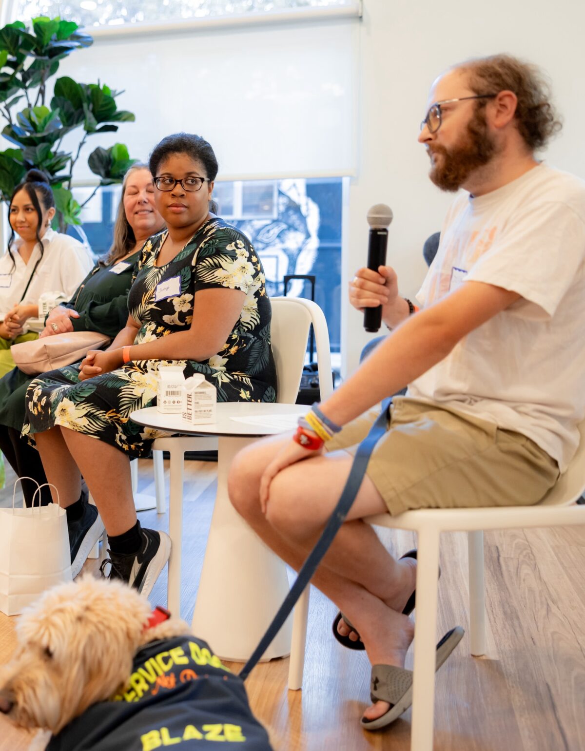 Four people sitting in chairs. One, a white man, is holding a microphone and a service dog sits next to him. Another, a black woman, is looking at him. They are in the lobby of The Kelsey Ayer Station.