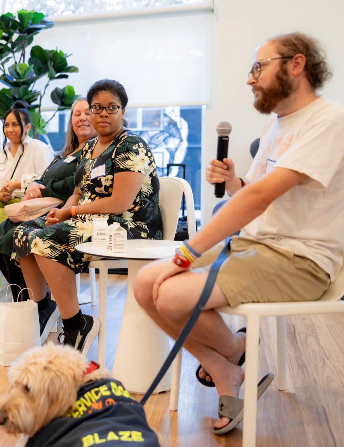 Four people sitting in chairs. One, a white man, is holding a microphone and a service dog sits next to him. Another, a black woman, is looking at him. They are in the lobby of The Kelsey Ayer Station.