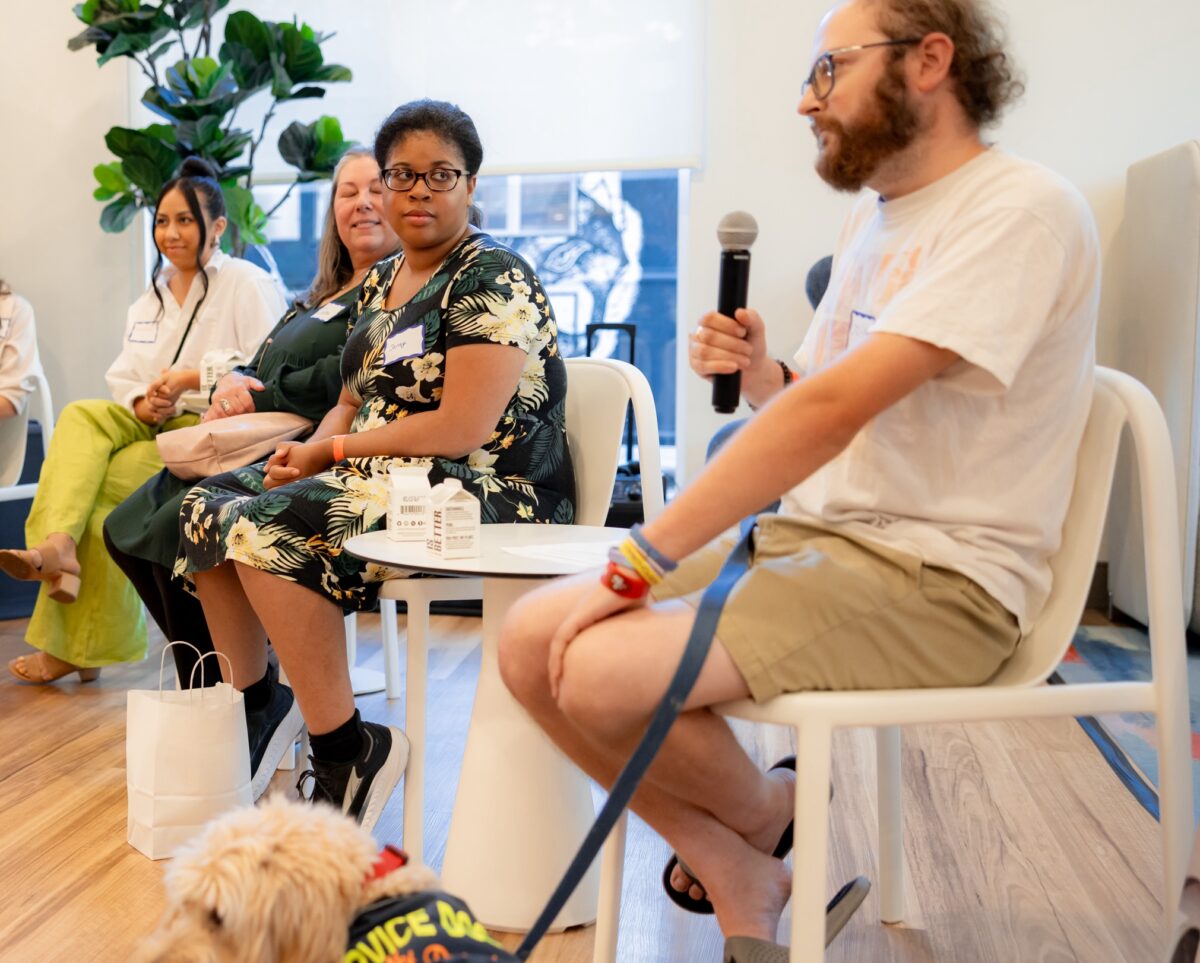 Four people sitting in chairs. One, a white man, is holding a microphone and a service dog sits next to him. Another, a black woman, is looking at him. They are in the lobby of The Kelsey Ayer Station.