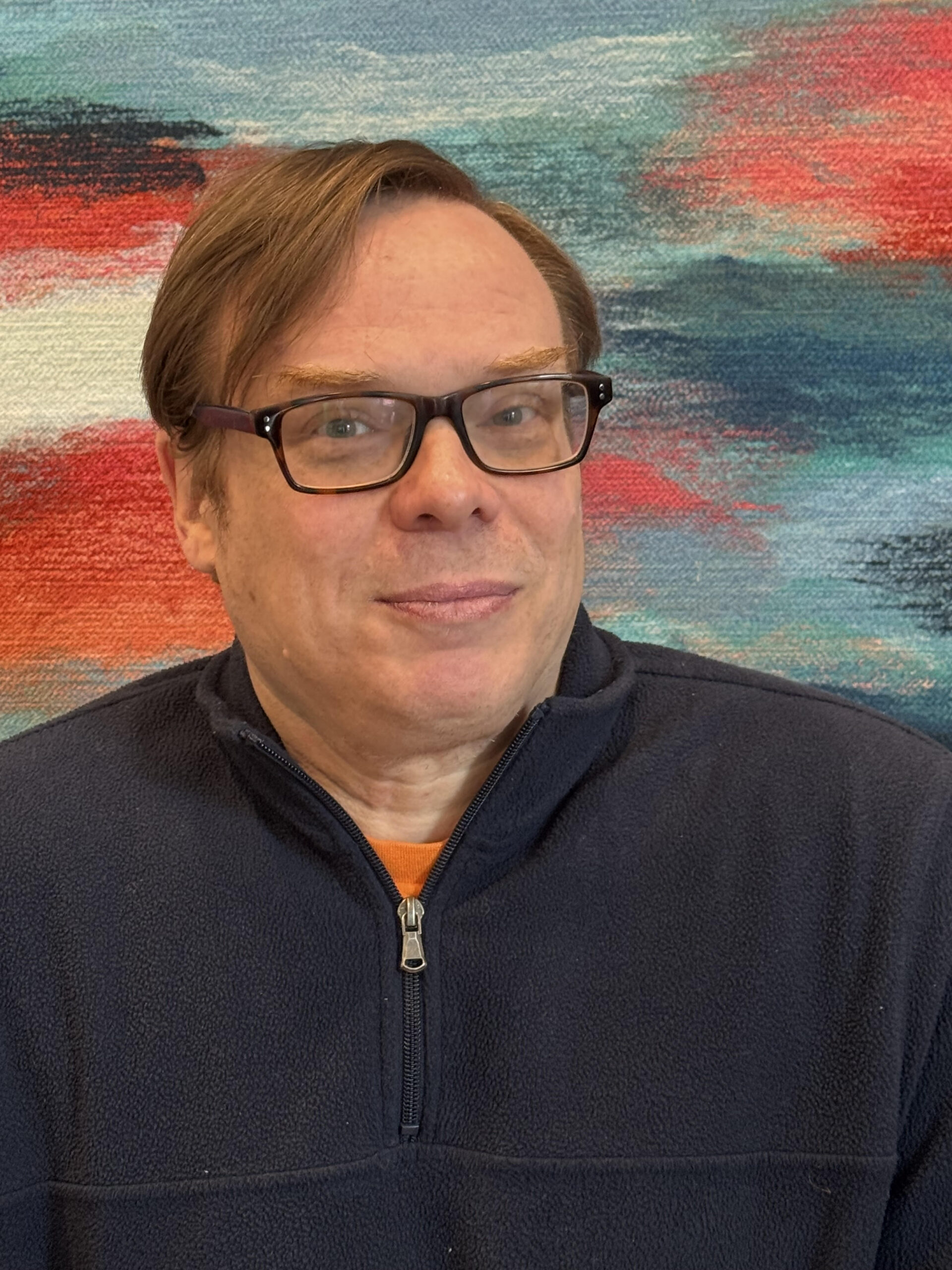 Doug is a light skinned man with red hair. He is wearing a dark blue pullover sweater with an orange shirt under it. He is also wearing dark-colored glasses. He is pictured against a multi-colored background.