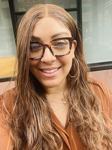 Ebony Salazar, a tan-skinned woman, with long light brown hair, wearing a brown shirt. She is wearing gold hoop earrings and brown glasses.