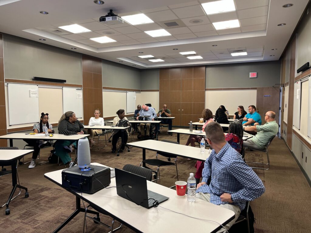 People sit at long tables in a conference room. Those sitting in the front of the room turn to listen to someone speak in the back.