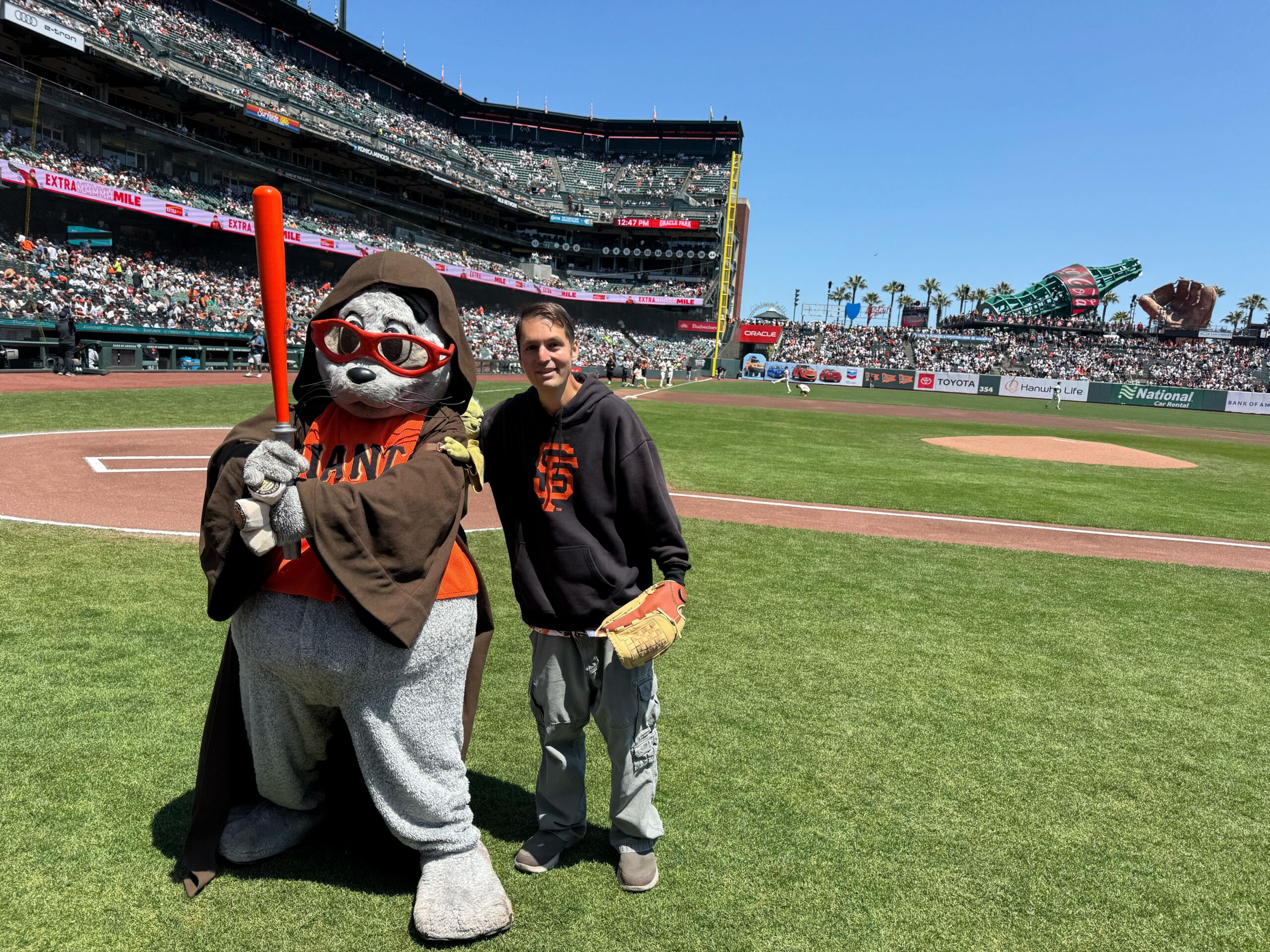 A resident poses with Lou Seal on the Giants baseball field. The resident is wearing a baseball glove, and Lou Seal is wearing a Star Wars cloak and holding an orange lightsaber.