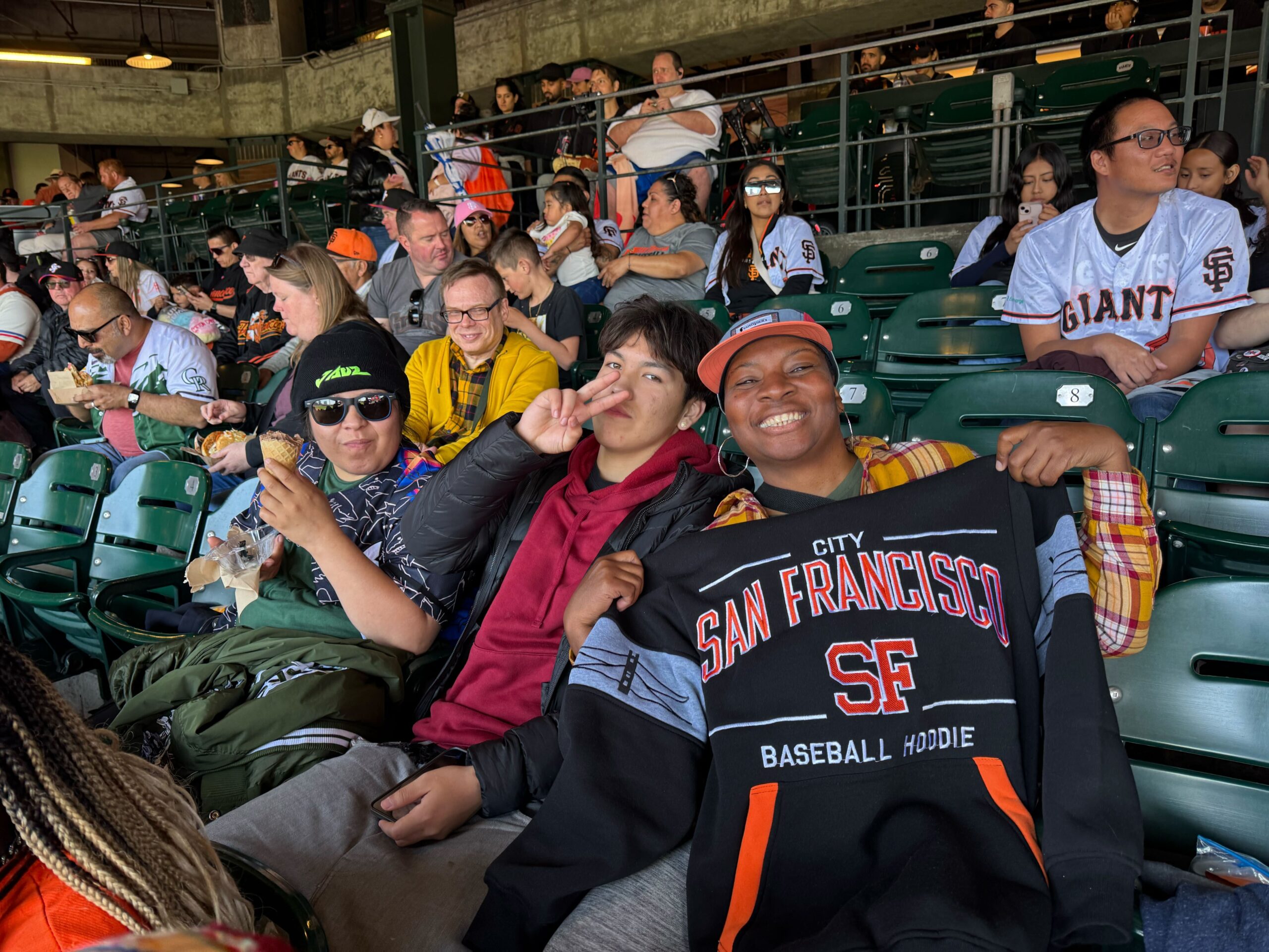 A group of residents of The Kelsey Ayer Station pose in the stands at a Giants game. One resident holds up a Giants’ sweatshirt and smiles.