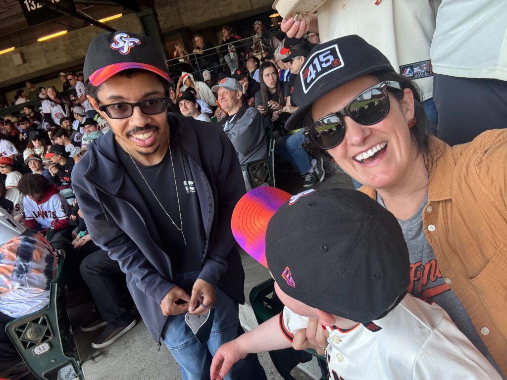 Isaac and Micaela, members of The Kelsey team, take a selfie in the stands of the Giants game. Micaela’s son sits on her lap. They are all wearing Giants baseball caps.