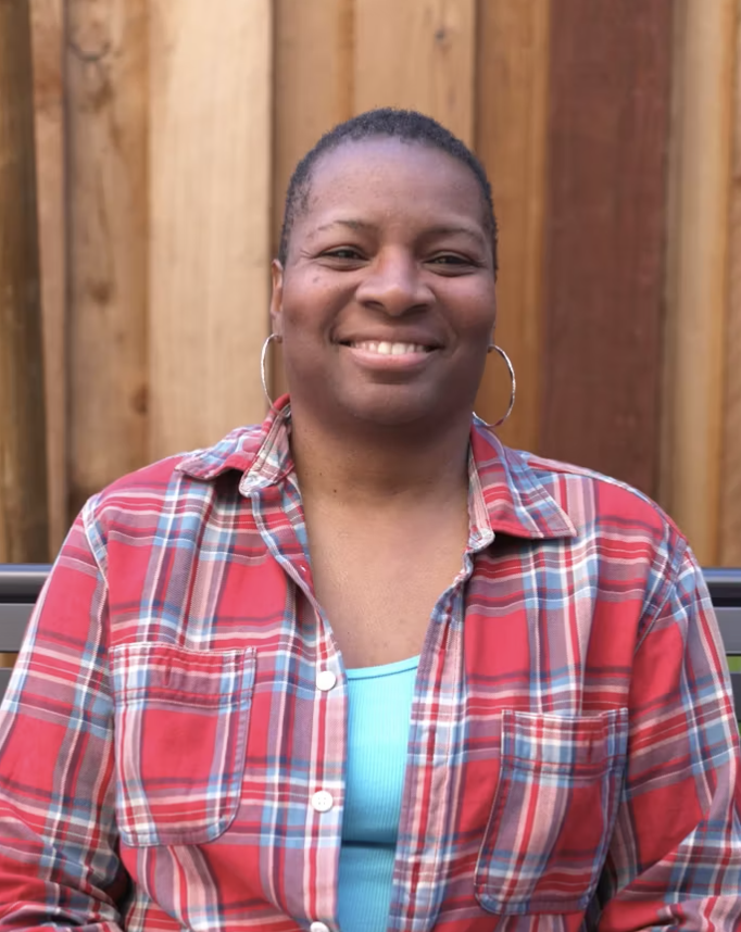 Carol, a Black woman with short dark hair, wearing a red and blue plaid collared shirt. She is smiling and sitting on a bench in front of a wood fence.