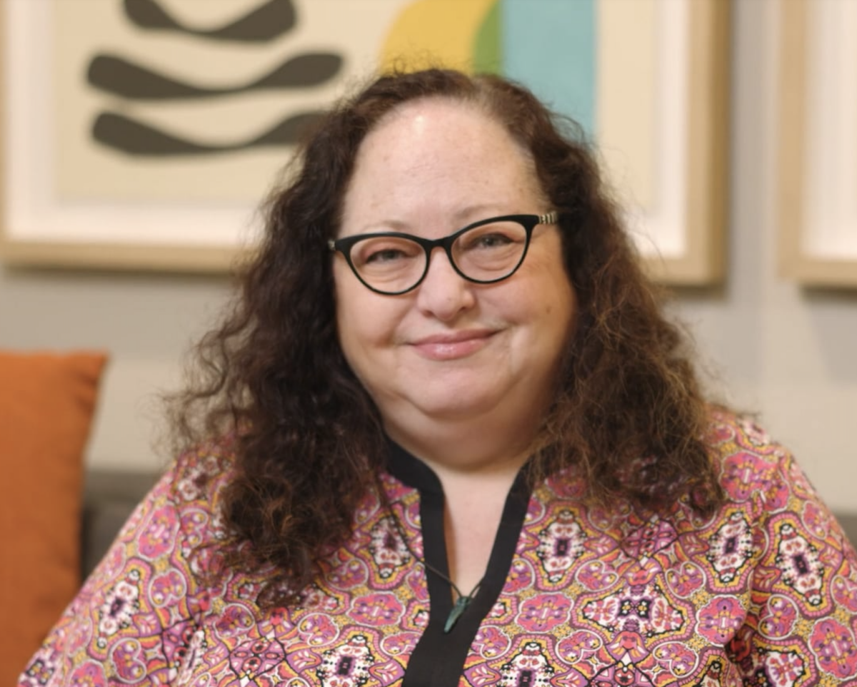 Sarah, a white woman with long curly hair and glasses, smiles at the camera. She wears a pink and purple patterned shirt.