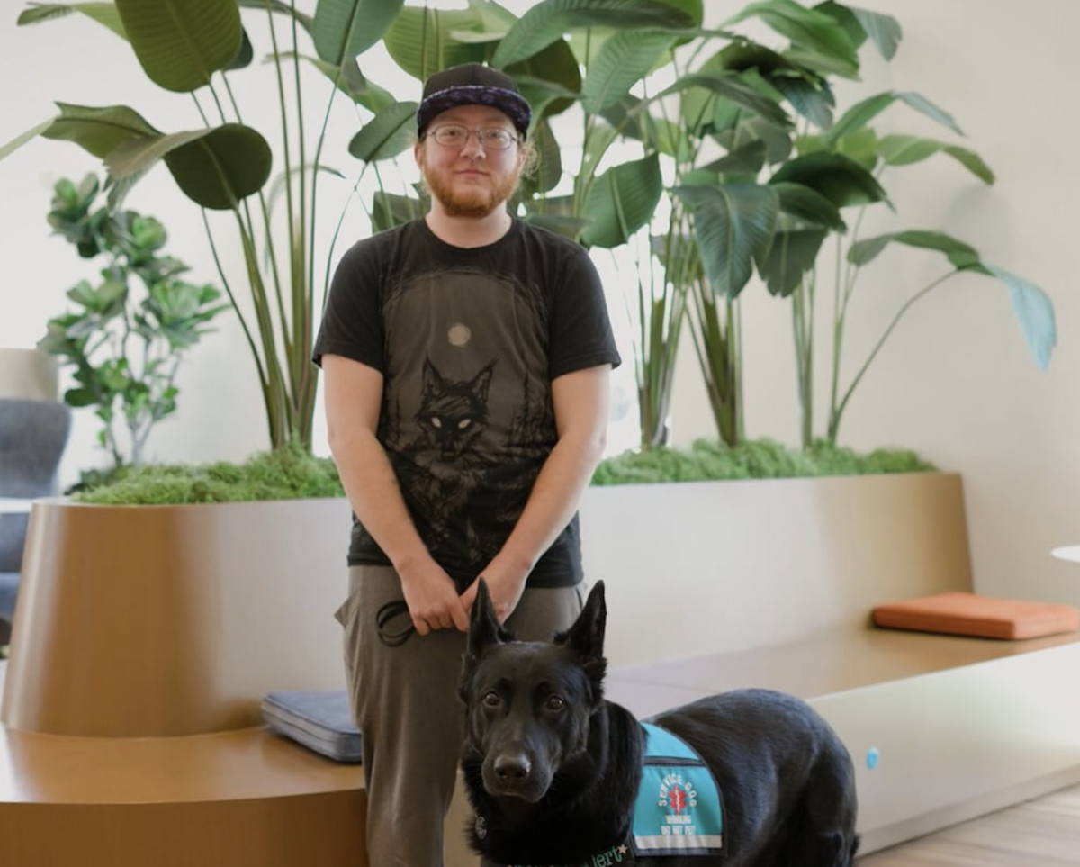 Tristan, a white man wearing glasses and a cap, stands with his service dog in the lobby of The Kelsey Ayer Station.