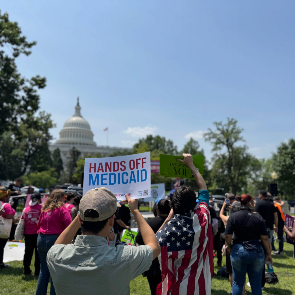 A man is pictured holding a sign that reads, “Hands Off Medicaid,” during a rally. A crowd is pictured, and the rotunda of the U.S. Capitol is visible in the background.