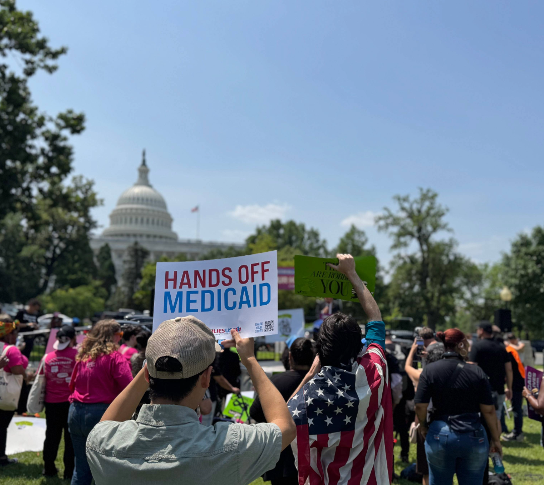A man is pictured holding a sign that reads, “Hands Off Medicaid,” during a rally. A crowd is pictured, and the rotunda of the U.S. Capitol is visible in the background.
