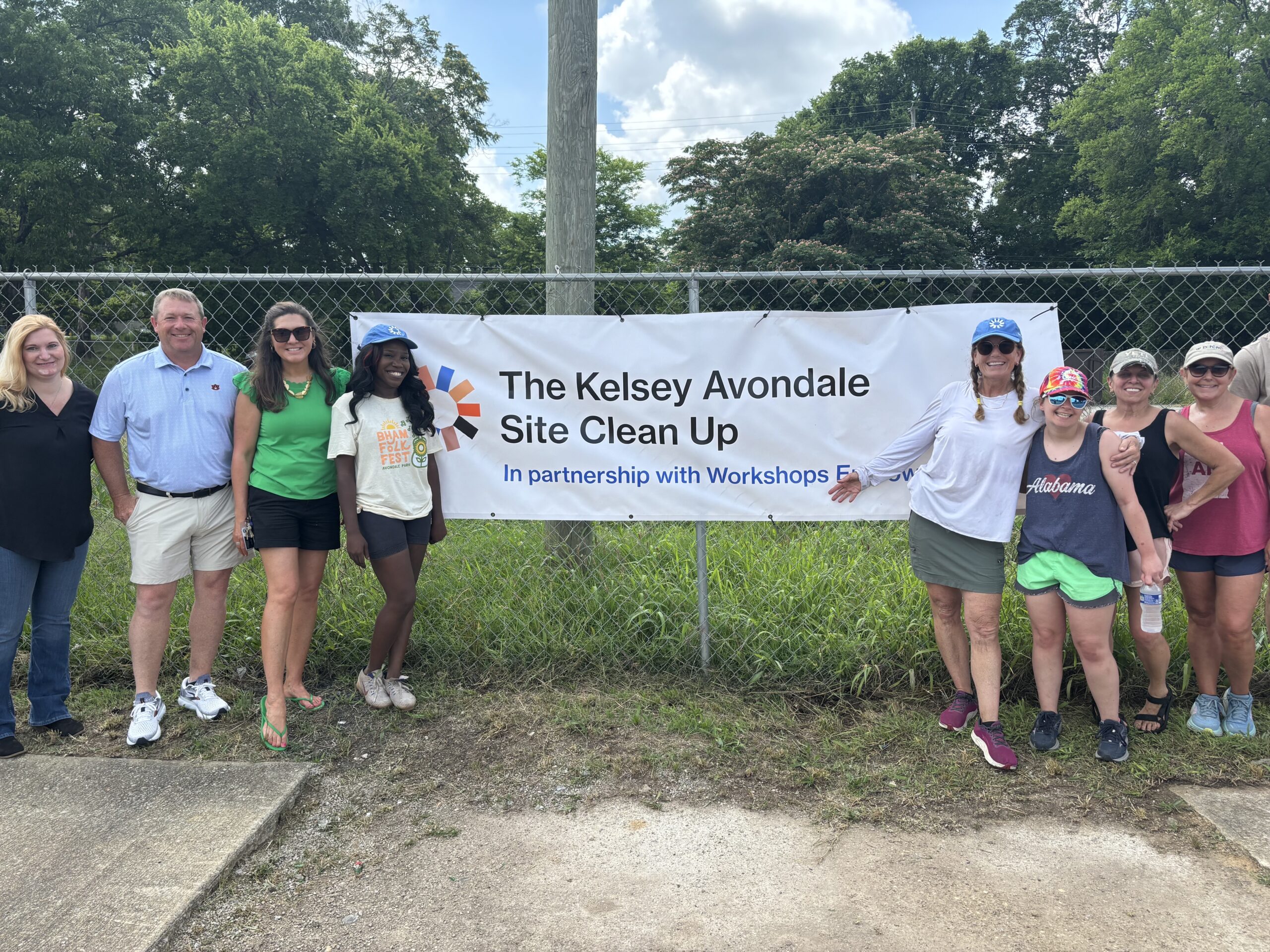 A group of people gather on both sides of a banner hanging on a fence. The banner reads, “The Kelsey Avondale Site Clean Up: In partnership with Workshops Empowerment.” The Kelsey’s logo is on the left side of the banner.