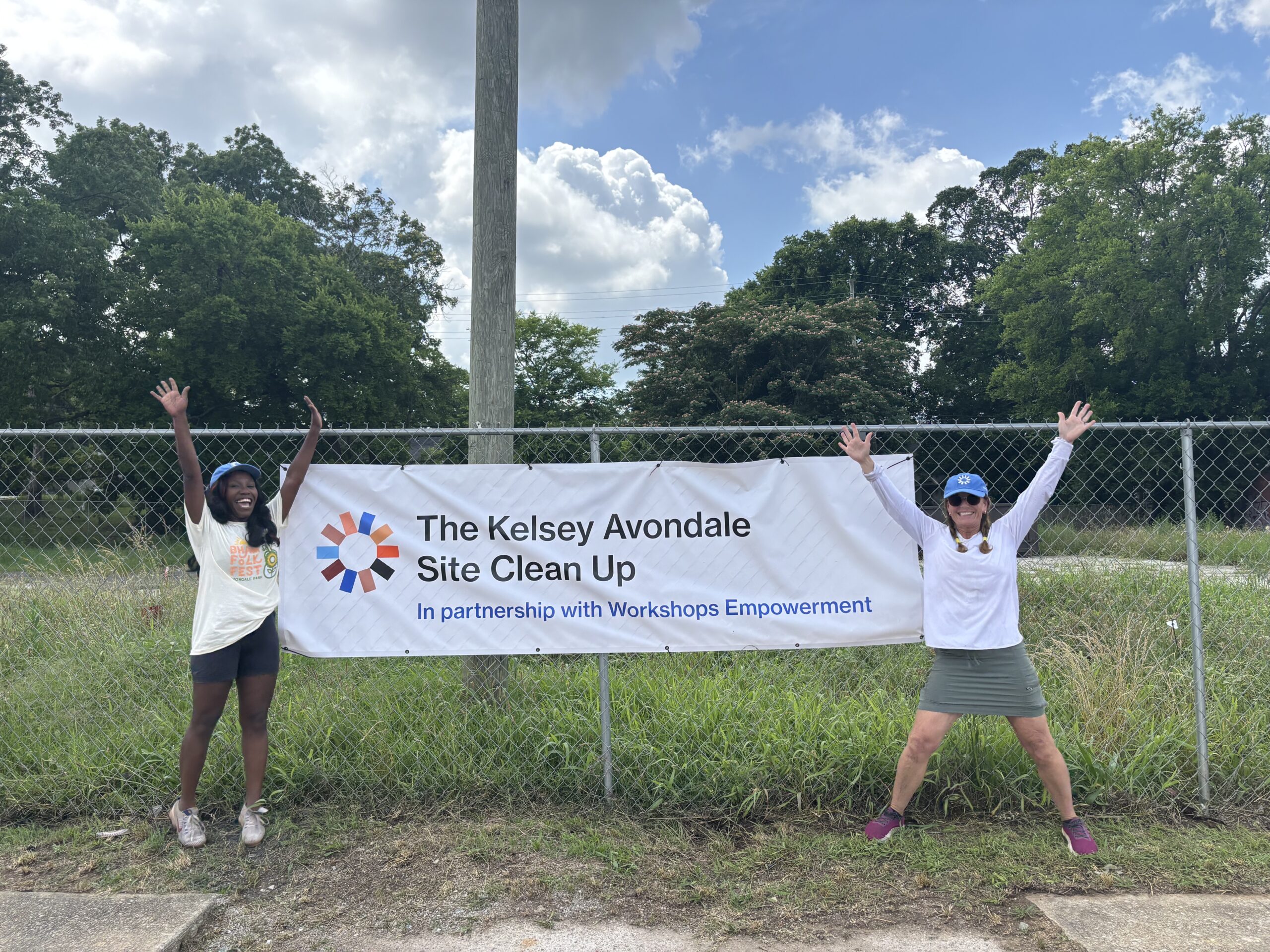 Brittney, The Kelsey’s Housing Manager, and Veronica, The Kelsey Avondale volunteer, pose with their hands in the air in front of The Kelsey Avondale site. They post on either side of The Kelsey Avondale Site Clean Up banner.