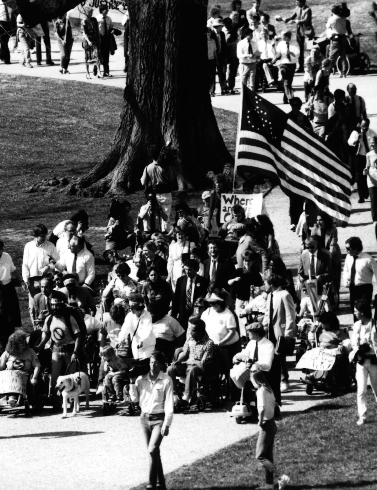 Protestors with and without disabilities march along a winding path. One holds a large American flag with the International Symbol of Access on it. This picture is from a 1990 demonstration led by Adapt in Washington, D.C., to support the Americans With Disabilities Act.