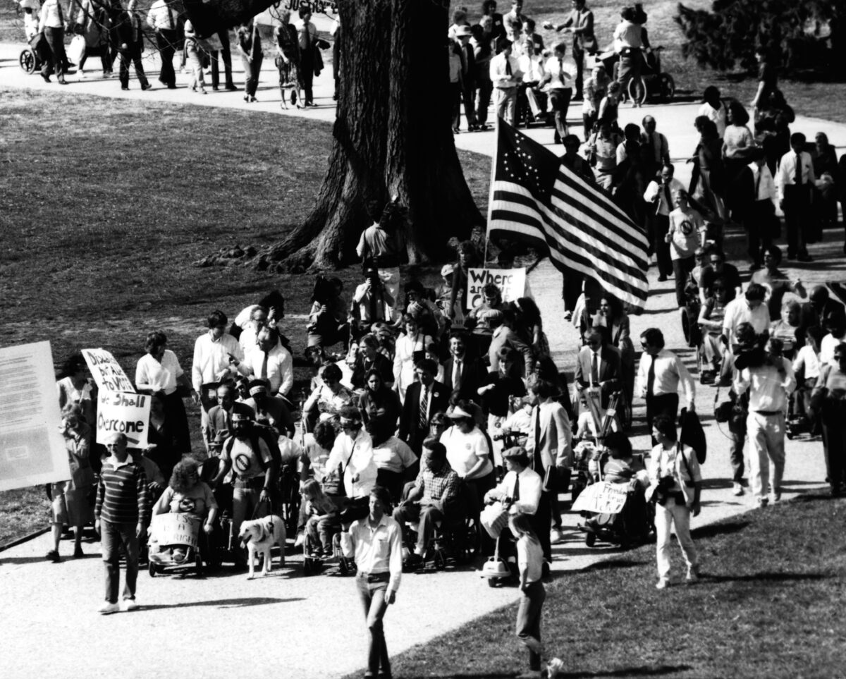 Protestors with and without disabilities march along a winding path. One holds a large American flag with the International Symbol of Access on it. This picture is from a 1990 demonstration led by Adapt in Washington, D.C., to support the Americans With Disabilities Act.