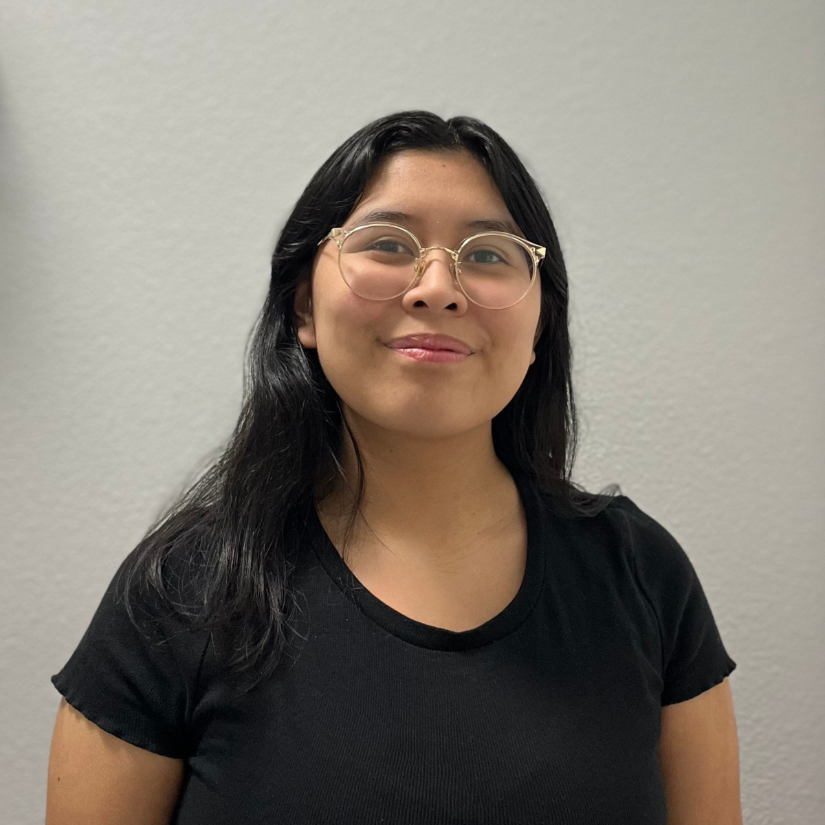 Image Description: Headshot of a Chicana woman with glasses wearing a short-sleeved black shirt with black medium-length hair smiling in front of a gray background.