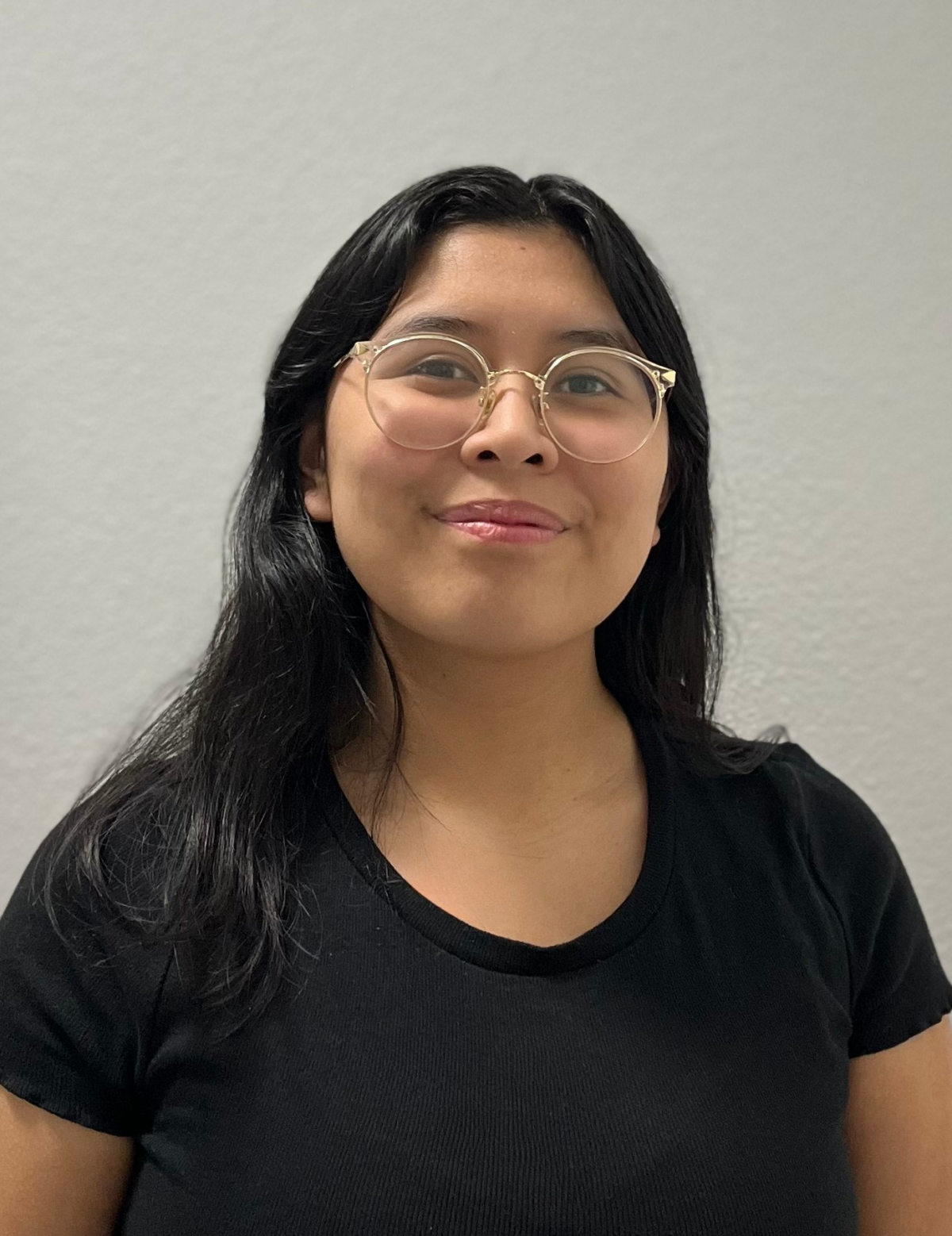 Headshot of a Chicana woman with glasses wearing a short-sleeved black shirt with black medium-length hair smiling in front of a gray background.