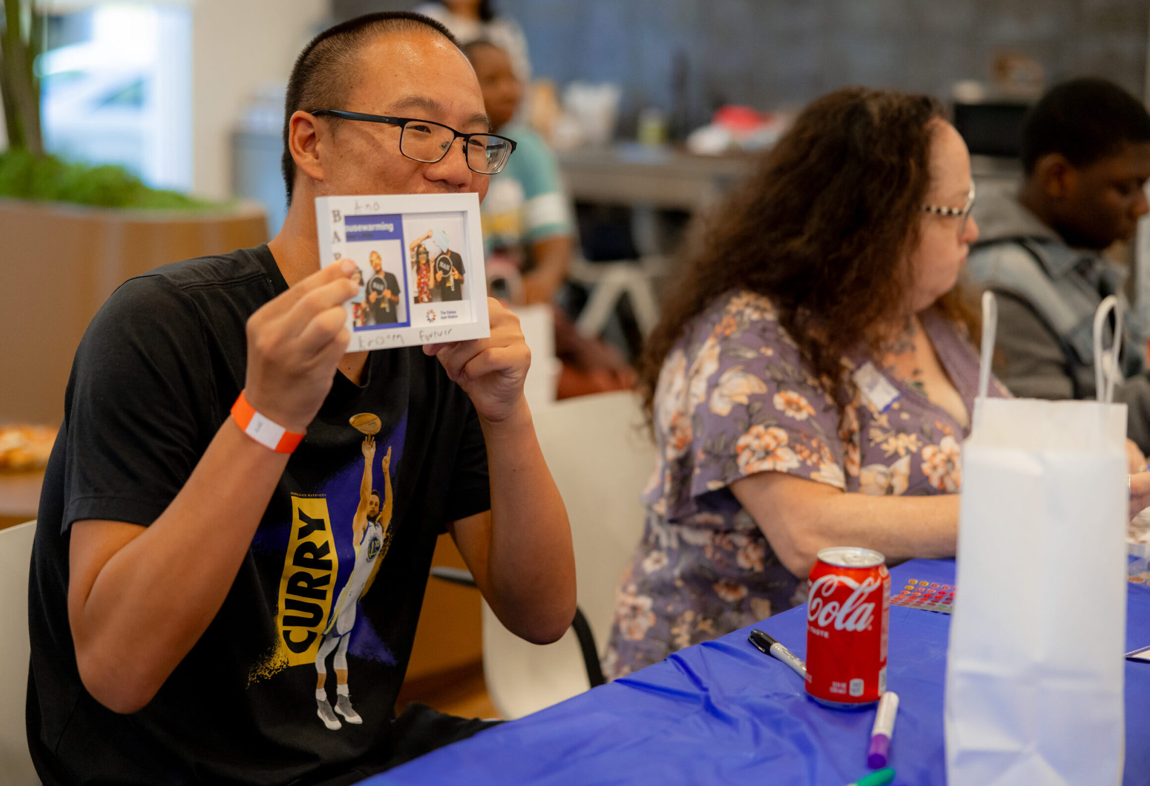 A resident smiles and holds a decorated picture frame with two photobooth images of him and other residents at The Kelsey Ayer Station Housewarming.