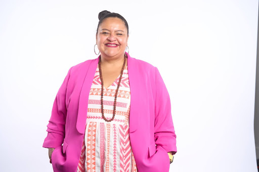 Image Description: Headshot of a caramel brown skin Mexican woman wearing a pink blazer and patterned dress, against a white background. She has large hoop earrings and a long beaded necklace. Her hair is styled up, and she’s smiling at the camera.