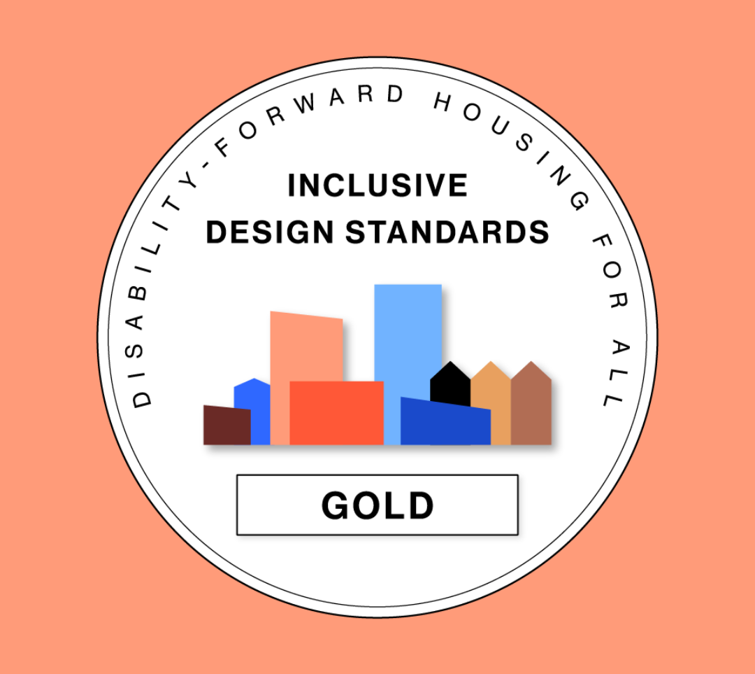A circular emblem with the words “Disability-forward housing for all” around the outer ring. In the center, bold text reads “Inclusive design standards.” Below the text is a row of colorful geometric building shapes in shades of red, orange, blue, and brown, representing a community skyline. Beneath the buildings is a rectangular box with the word “Gold” in bold black letters, signifying the project’s certification level. The background is coral.