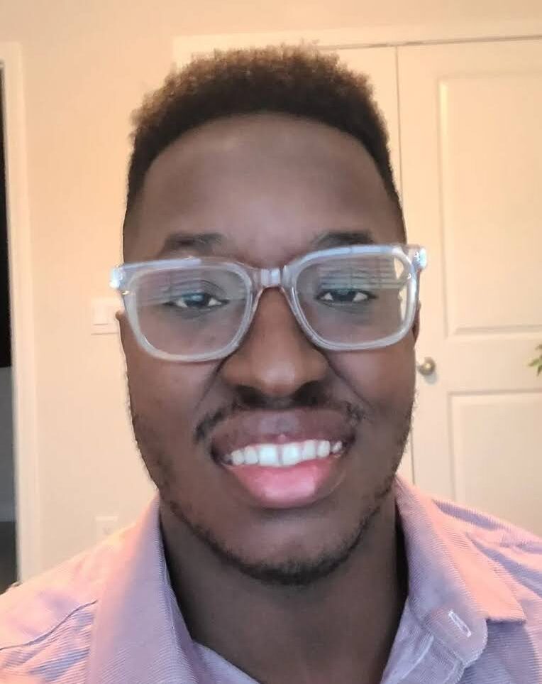 Image description: Chijioke, a Black, dark-skinned man wearing a pinkish red striped dress shirt standing with a door in the background.