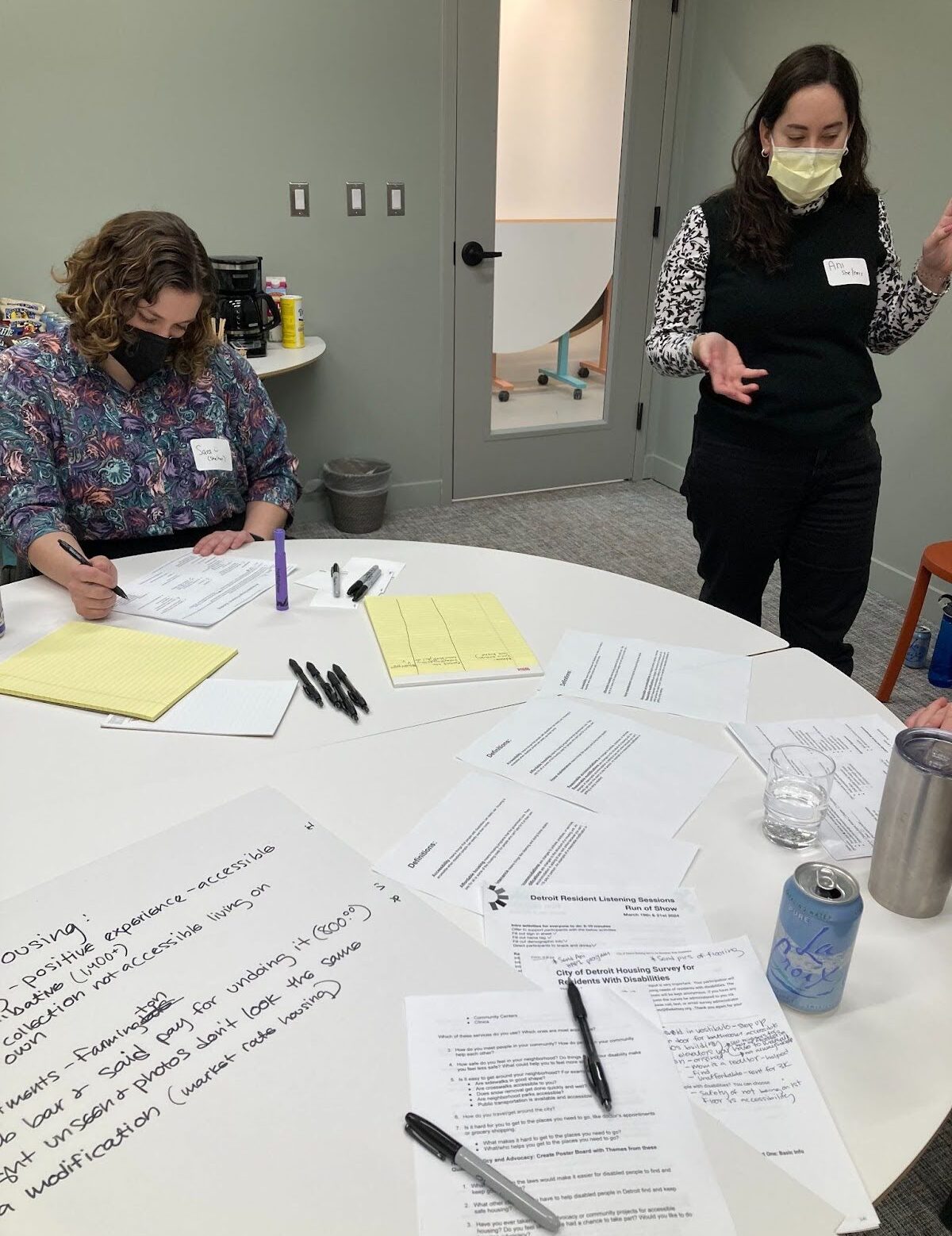 Two people are pictured in a meeting room wearing masks. One is sitting and writing at a table strewn with papers and pens, and the other is standing and talking. A glass door is visible behind them.