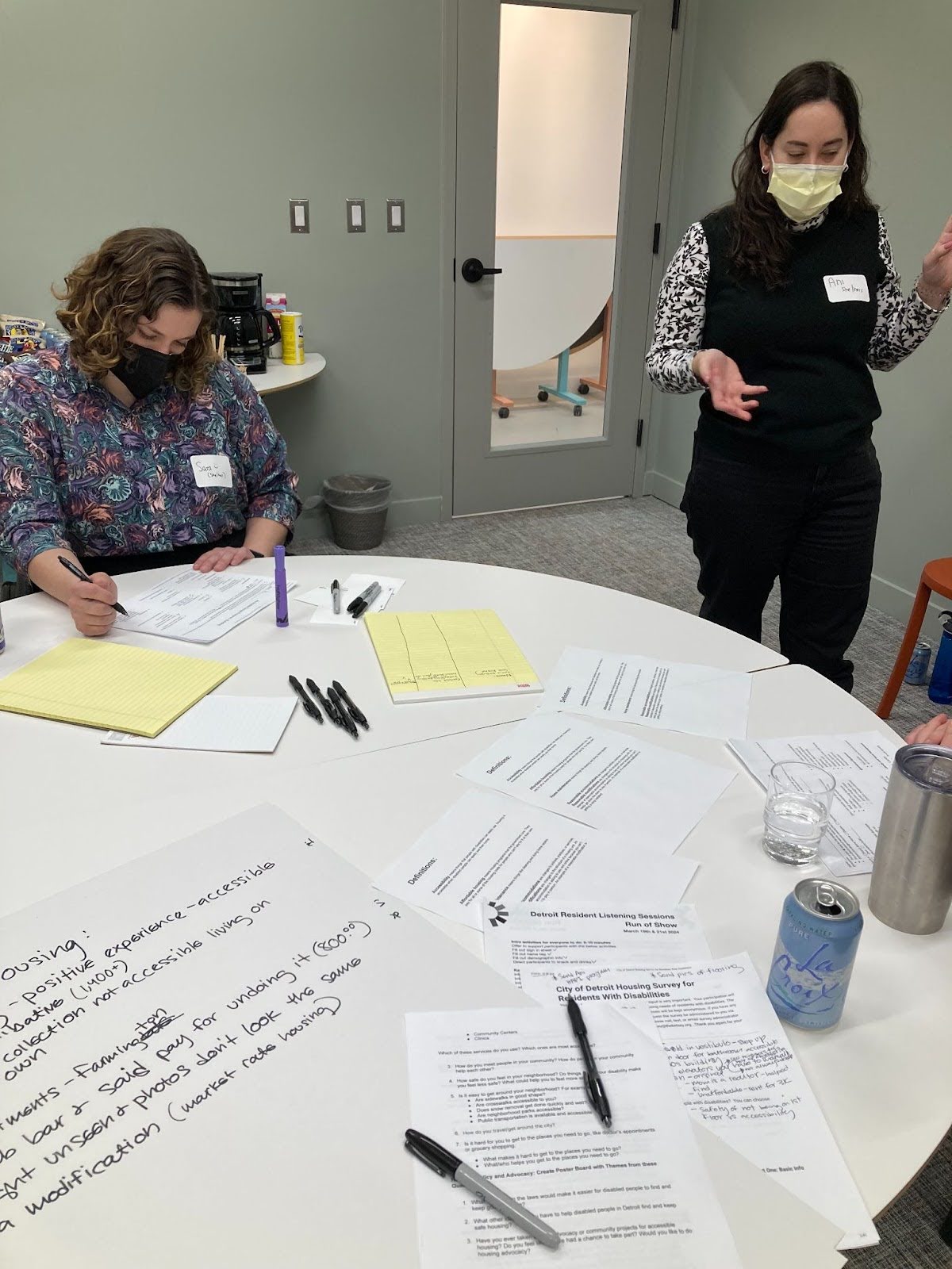 Two people are pictured in a meeting room wearing masks. One is sitting and writing at a table strewn with papers and pens, and the other is standing and talking. A glass door is visible behind them.