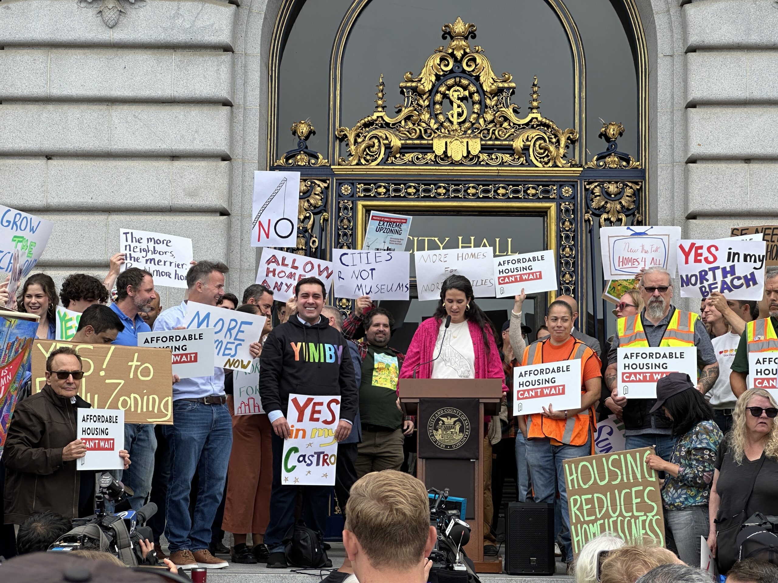 A rally at San Francisco City Hall. People stand with various paper and cardboard signs that read things like, “Affordable housing can’t wait,” “Housing reduces homelessness,” and “Yes in my Castro.” In the middle, Micaela Connery, The Kelsey’s CEO and co-founder, gives remarks at a podium with a microphone. She is a tall, white woman with long brown hair wearing a hot pink cardigan and a graphic tee with the shape of California on it.