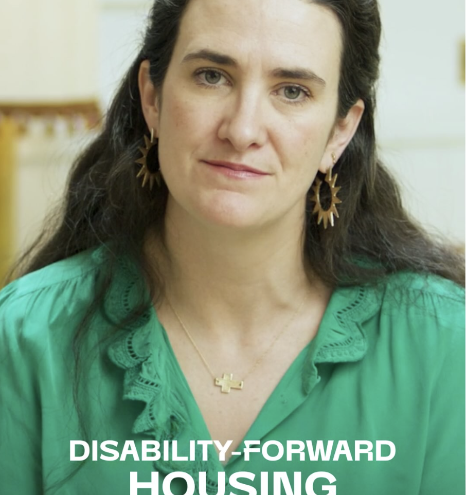 A still image from Micaela Connery’s Brief But Spectacular segment. Micaela looks at the camera with a slight smile. She is a white woman with long, brown hair, green eyes, wearing a green blouse and gold jewelry, including a cross necklace and sun-shaped earrings. The text “Disability-Forward Housing: Micaela Connery,” is in the lower third of the image.