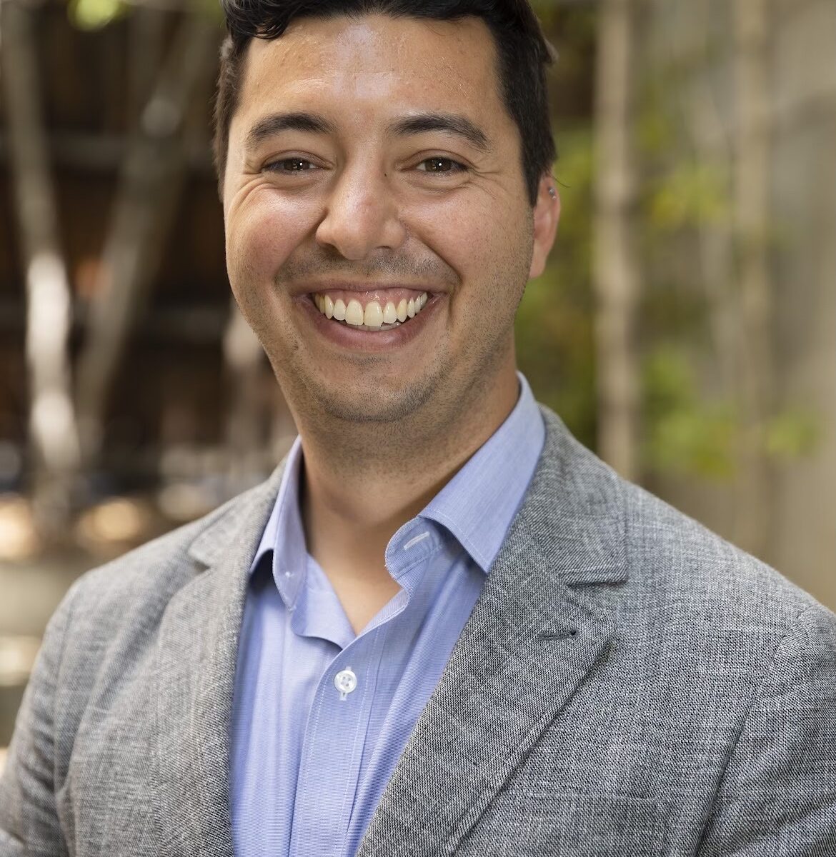 Sam, a white, dark-haired man wearing a gray blazer and light blue shirt, is standing outdoors with trees in the background.
