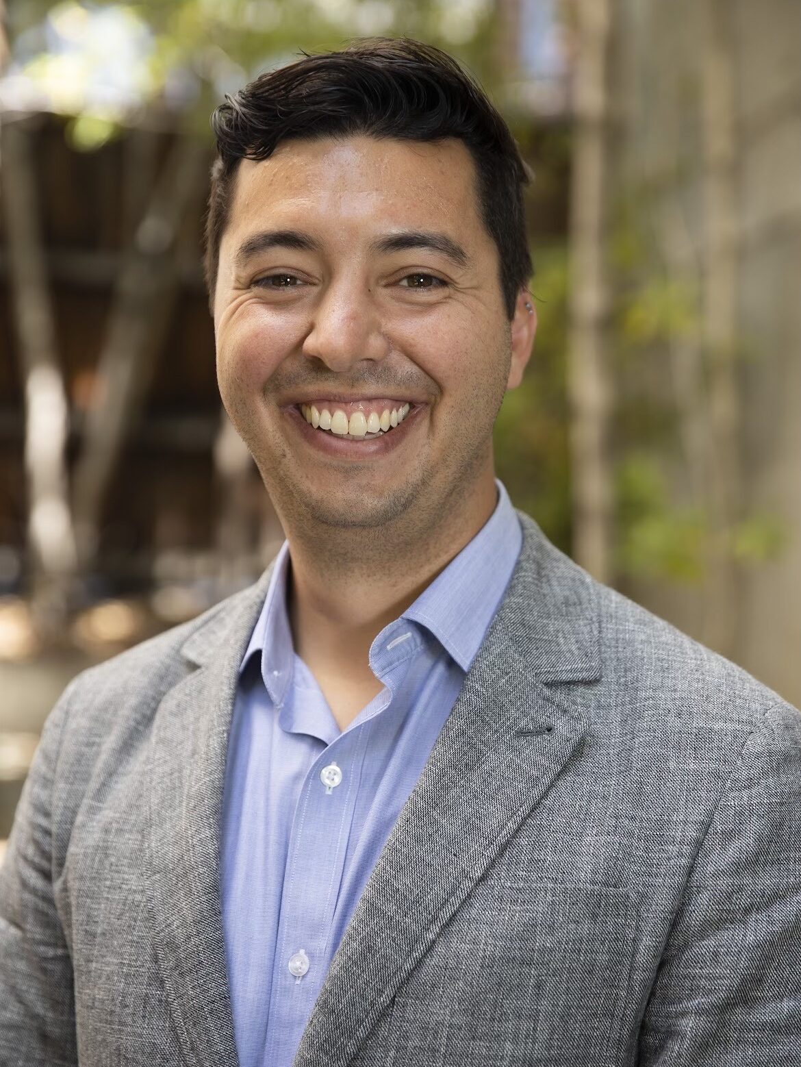 Sam, a white, dark-haired man wearing a gray blazer and light blue shirt, is standing outdoors with trees in the background.