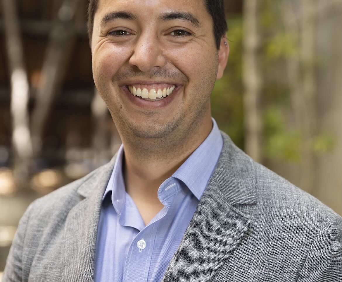 Sam, a white, dark-haired man wearing a gray blazer and light blue shirt, is standing outdoors with trees in the background.