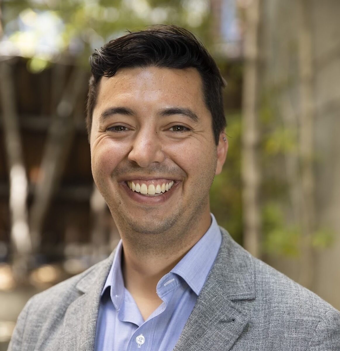 Sam, a white, dark-haired man wearing a gray blazer and light blue shirt, is standing outdoors with trees in the background.
