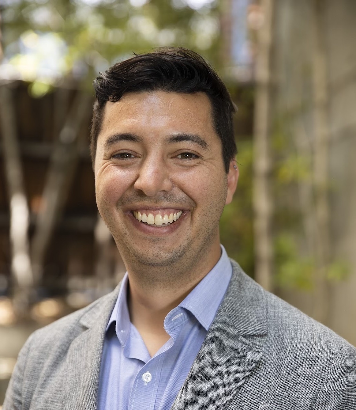 Sam, a white, dark-haired man wearing a gray blazer and light blue shirt, is standing outdoors with trees in the background.