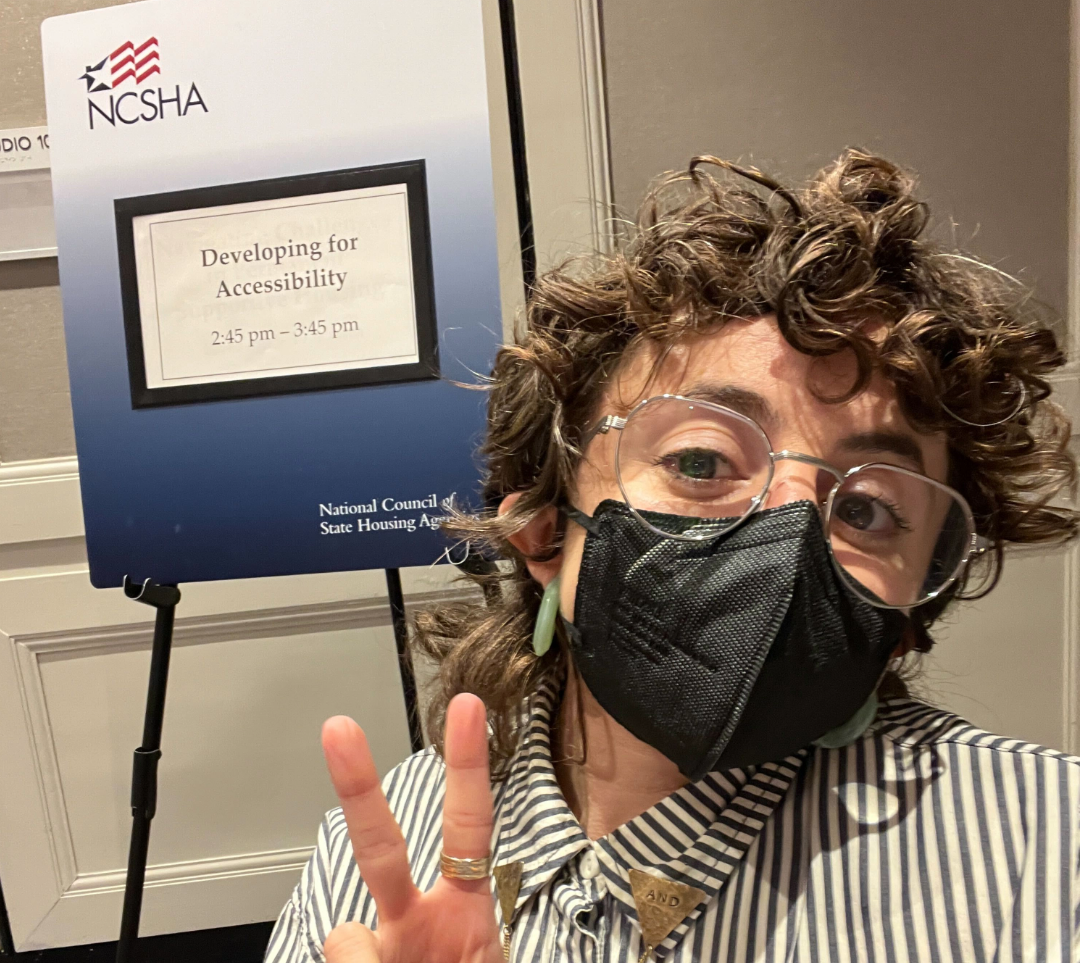 Allie is a white queer person with glasses, wearing a black and white striped shirt and a black mask. Allie is pictured in front of an easel with a sign on it with the logo of the National Council for State Housing Agencies. The sign says the name of their session, “Developing for Accessibility, 2:45pm-3:45 pm.”