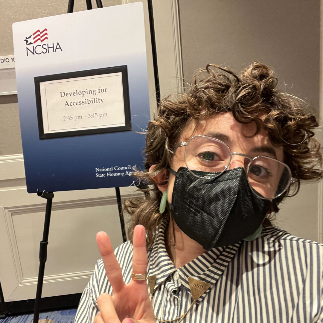 Allie is a white queer person with glasses, wearing a black and white striped shirt and a black mask. Allie is pictured in front of an easel with a sign on it with the logo of the National Council for State Housing Agencies. The sign says the name of their session, “Developing for Accessibility, 2:45pm-3:45 pm.”