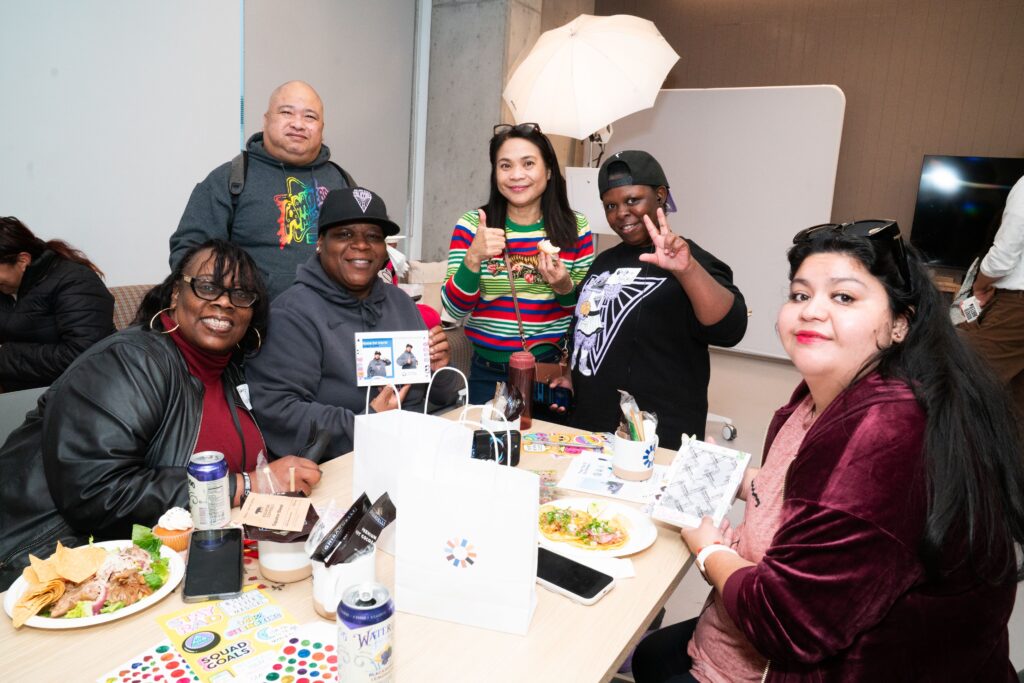 Image Description: Residents and guests gather sitting and standing at a table in a bright community room. There are bags, food, and stickers covering the table.