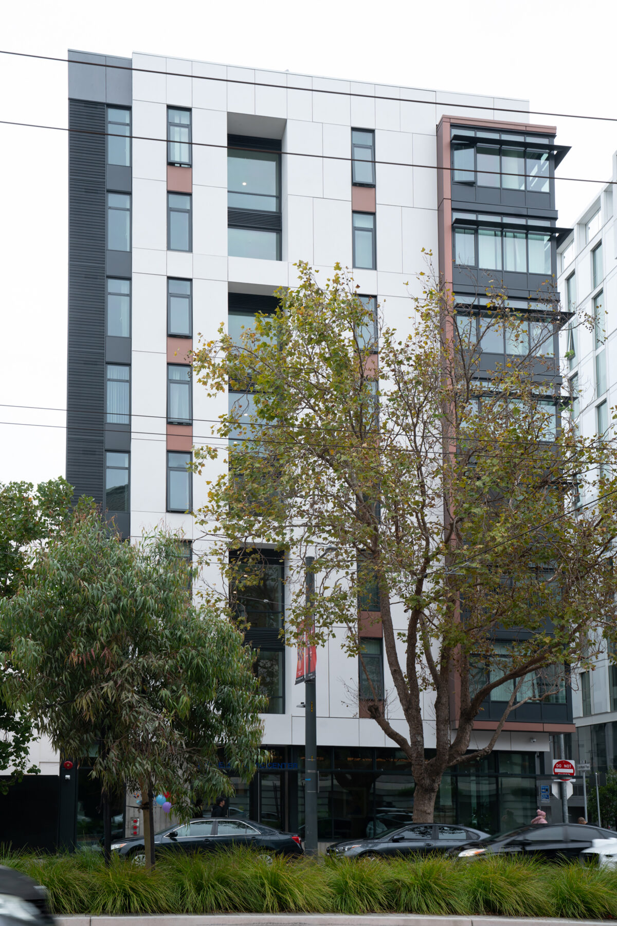 The exterior of The Kelsey Civic Center, a modern building with a white, black, and brown facade and large windows. Greenery, street signage, and traffic are visible on the building's ground floor.