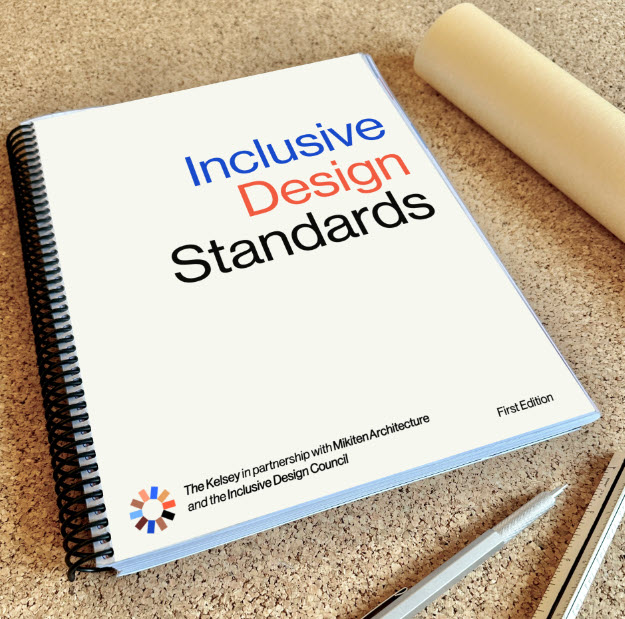 A spiral-bound book with “Inclusive Design Standards” on the cover rests on a corkboard along with a pen, a ruler, and some rolled-up paper.