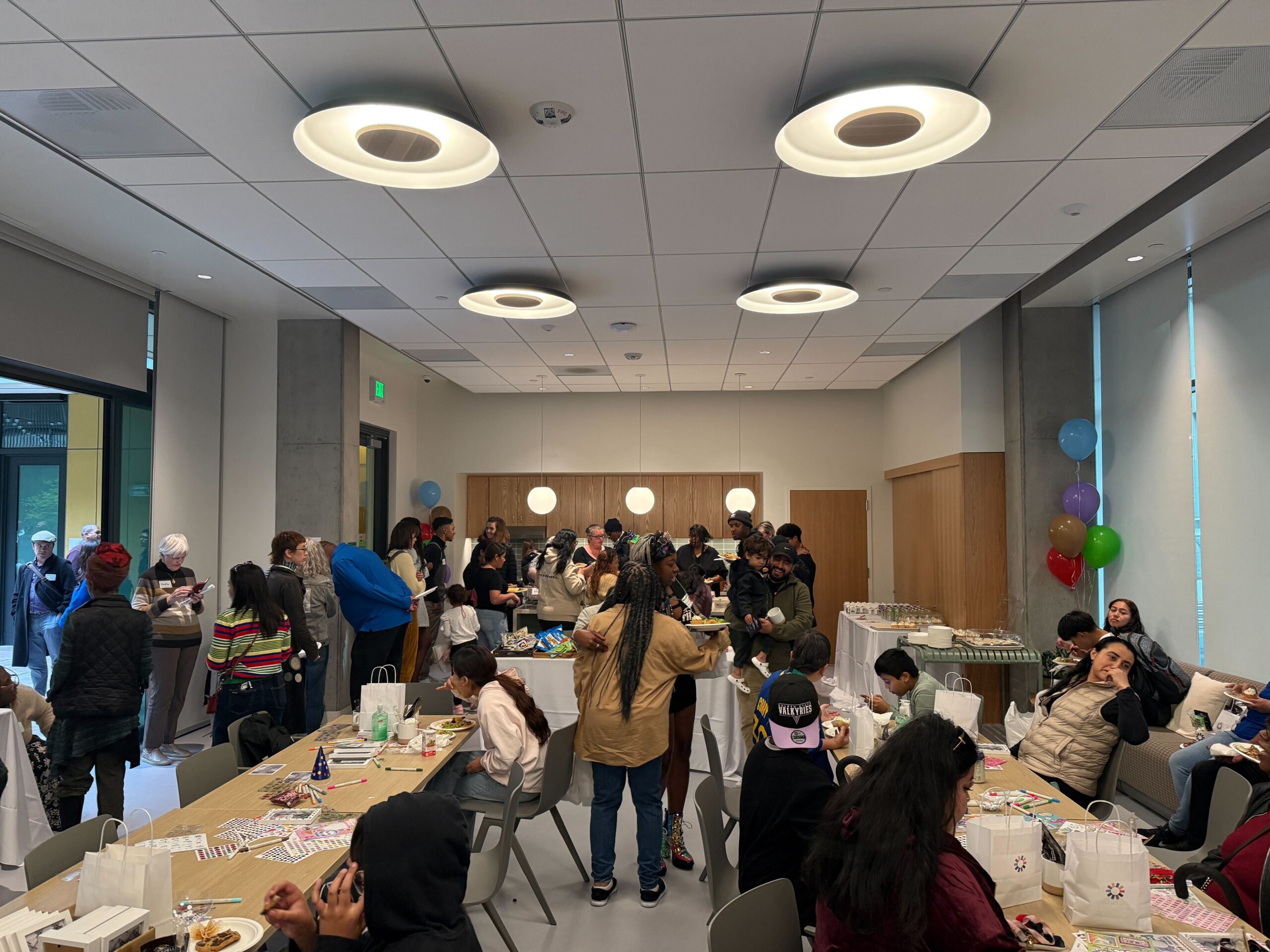 Residents and guests gather in a bright community room at The Kelsey Civic Center, enjoying food, conversation, and activities around long tables decorated with gift bags and balloons during the housewarming celebration.