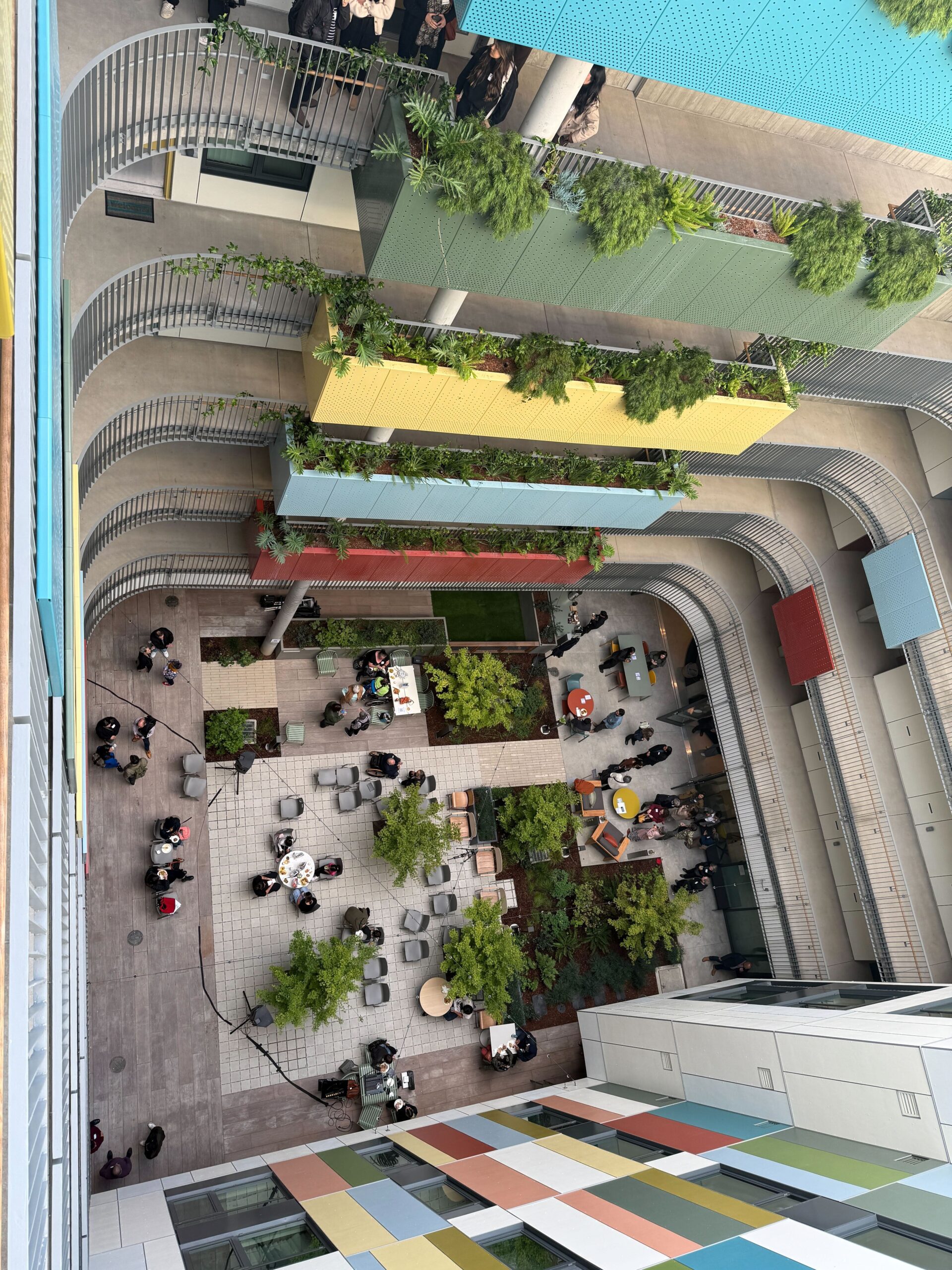 Aerial view of The Kelsey Civic Center’s colorful courtyard, where guests gather at tables among trees and plants during the housewarming celebration. The courtyard is surrounded by multicolored balconies with greenery. Guests can be seen on one of the balconies, taking a tour of the building.