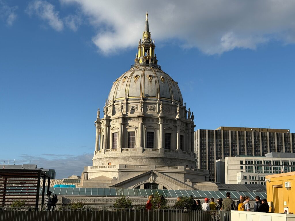 View of San Francisco City Hall’s grand dome under a blue sky, seen from The Kelsey Civic Center rooftop where guests gather along the terrace during the housewarming celebration.